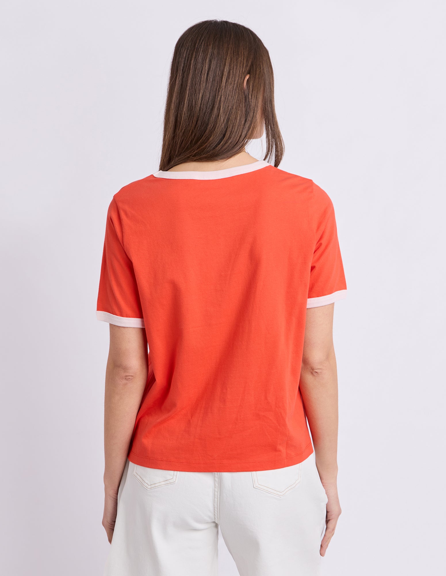 A woman viewed from the back wearing a bright orange short-sleeve ringer T-shirt with white trim at the collar and sleeve cuffs, paired with white pants, against a light gray studio background.