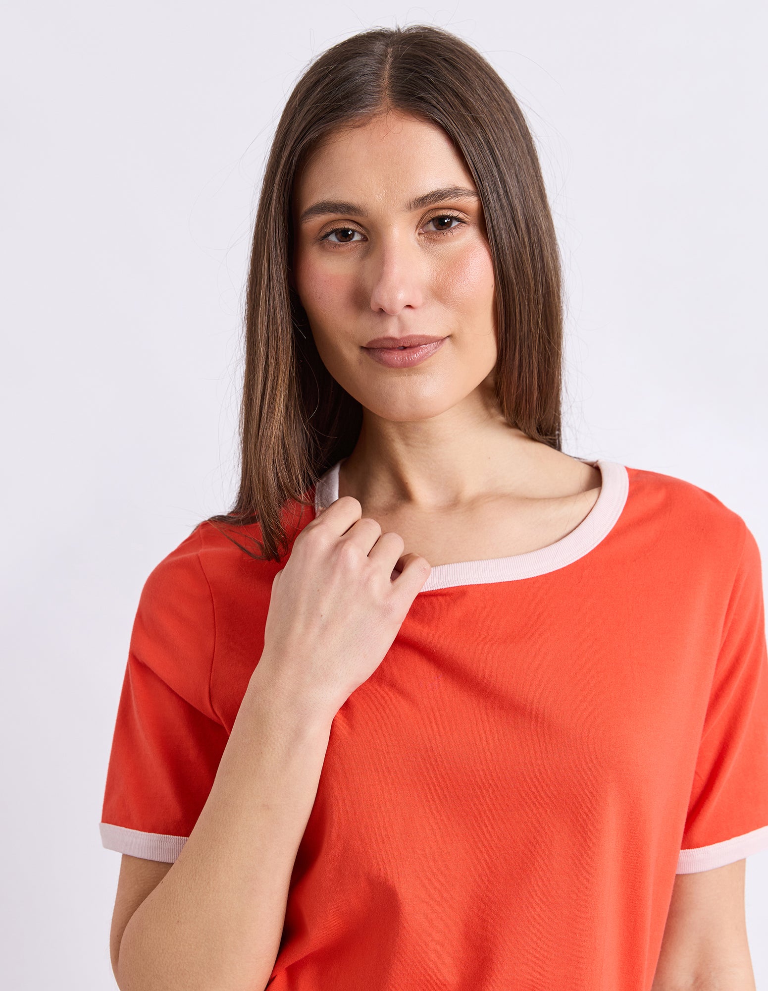 A woman with long brown hair wearing a bright orange short-sleeve t-shirt with a contrasting light pink crew neck and sleeve cuffs, set against a plain light gray background.