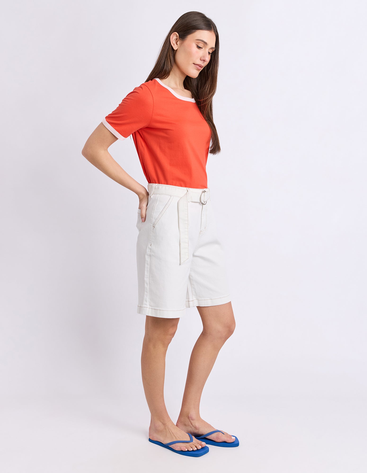 A woman modeling a bright orange short-sleeve t-shirt with white ringer trim, paired with off-white shorts and blue flip-flops, against a plain white studio background.