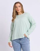 A woman modeling a sage green long-sleeve crewneck top with a relaxed, slightly cropped fit, paired with light blue jeans against a light gray studio background.