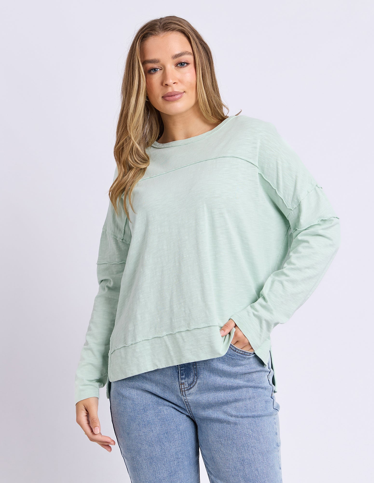 A woman modeling a sage green long-sleeve crewneck top with a relaxed, slightly cropped fit, paired with light blue jeans against a light gray studio background.