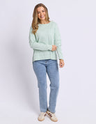 A woman modeling a sage green long-sleeve knit top with decorative layered sleeve cuffs, paired with light blue jeans and beige sneakers, against a plain white studio background.