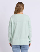 A woman viewed from the back wearing a light sage green long-sleeve top with horizontal seam details and light blue jeans, set against a plain light gray studio background.