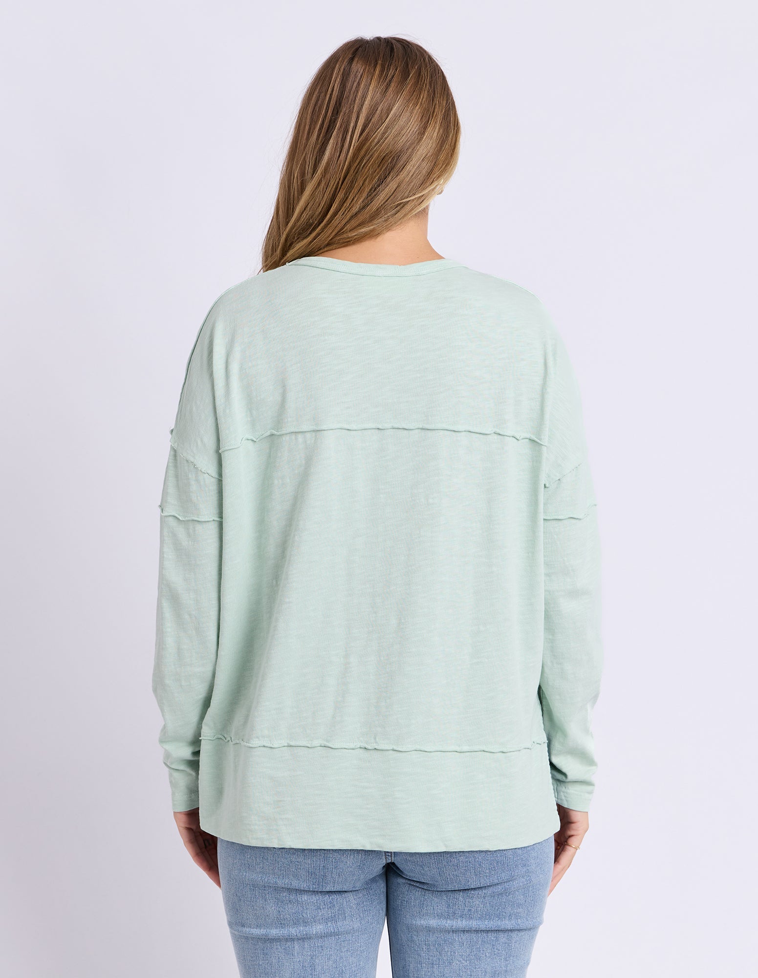 A woman viewed from the back wearing a light sage green long-sleeve top with horizontal seam details and light blue jeans, set against a plain light gray studio background.