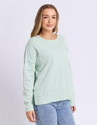 A woman modeling a light mint green long-sleeve crewneck sweatshirt with horizontal seam details, paired with blue jeans, against a light gray studio background.