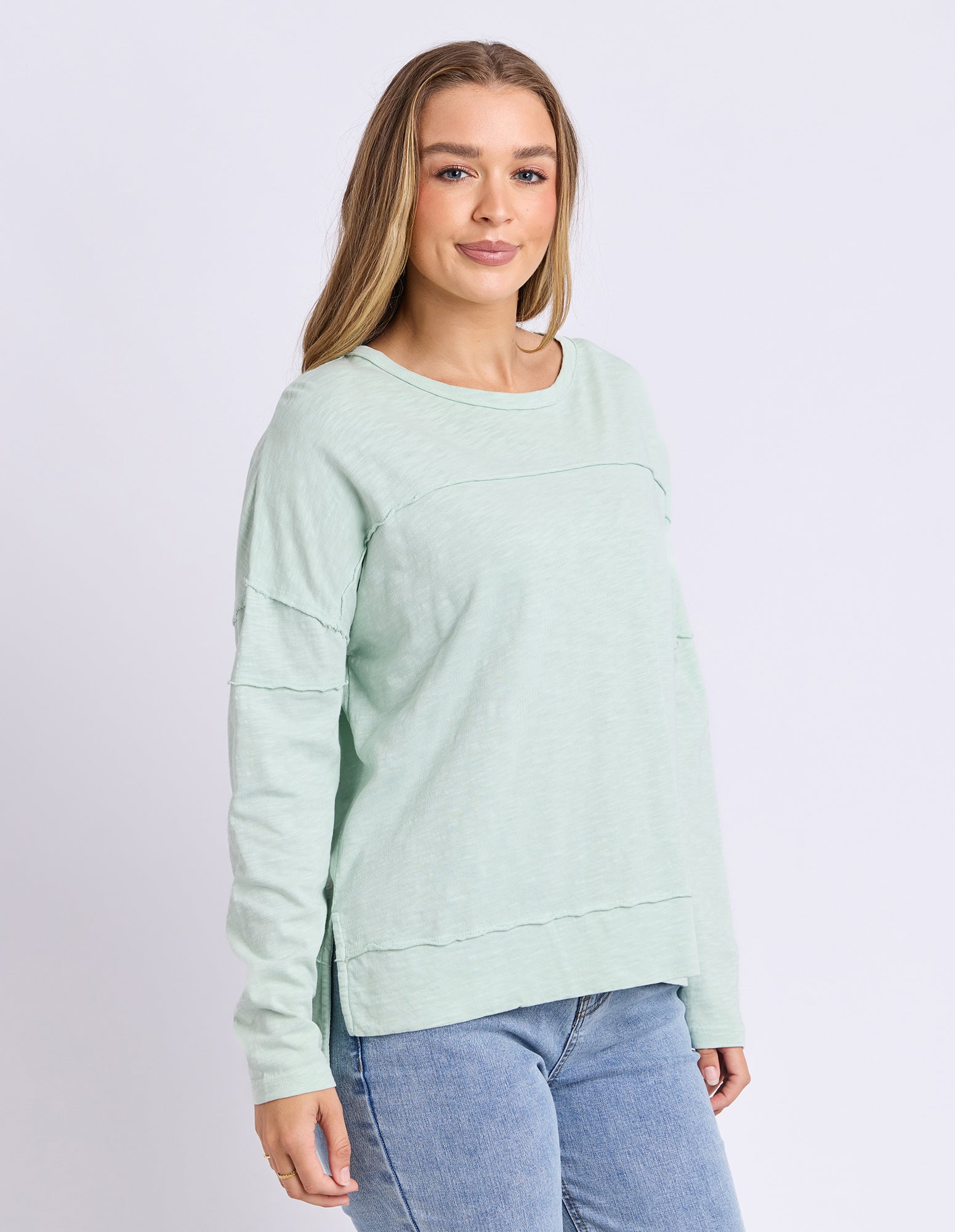 A woman modeling a light mint green long-sleeve crewneck sweatshirt with horizontal seam details, paired with blue jeans, against a light gray studio background.