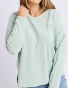A woman wearing a light mint green long-sleeve top with layered ruffle details on the chest and sleeves, paired with blue jeans against a light gray studio background.