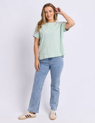 A woman modeling a light sage green short-sleeve crewneck sweatshirt and light-wash blue jeans, paired with beige sneakers, against a plain light gray studio background.