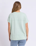 A woman viewed from the back wearing a light mint green short-sleeve top and blue jeans against a plain light background.