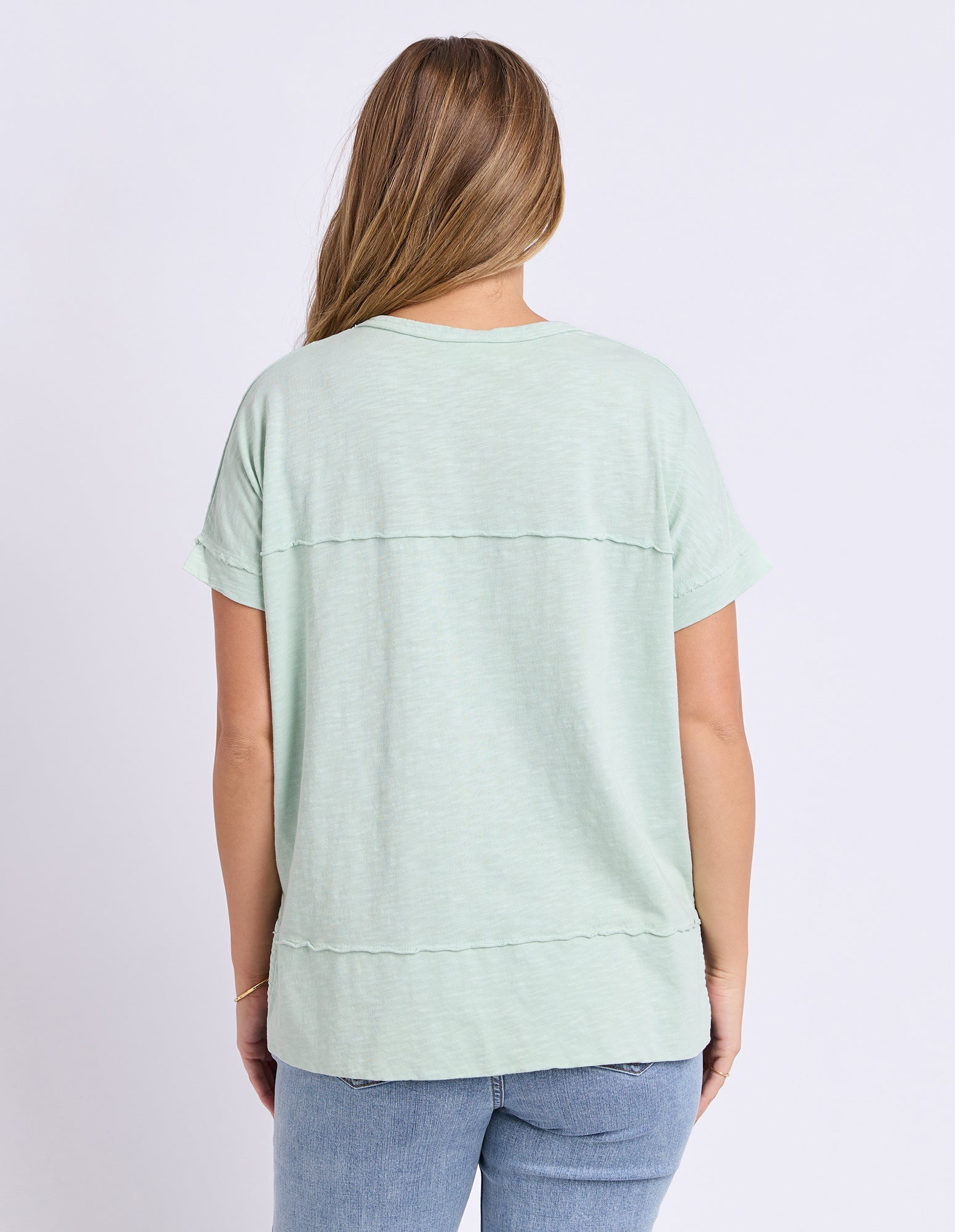 A woman viewed from the back wearing a light mint green short-sleeve top and blue jeans against a plain light background.
