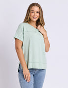 A woman modeling a light sage green short-sleeve top with a split neckline, paired with blue jeans, against a plain light gray studio background.