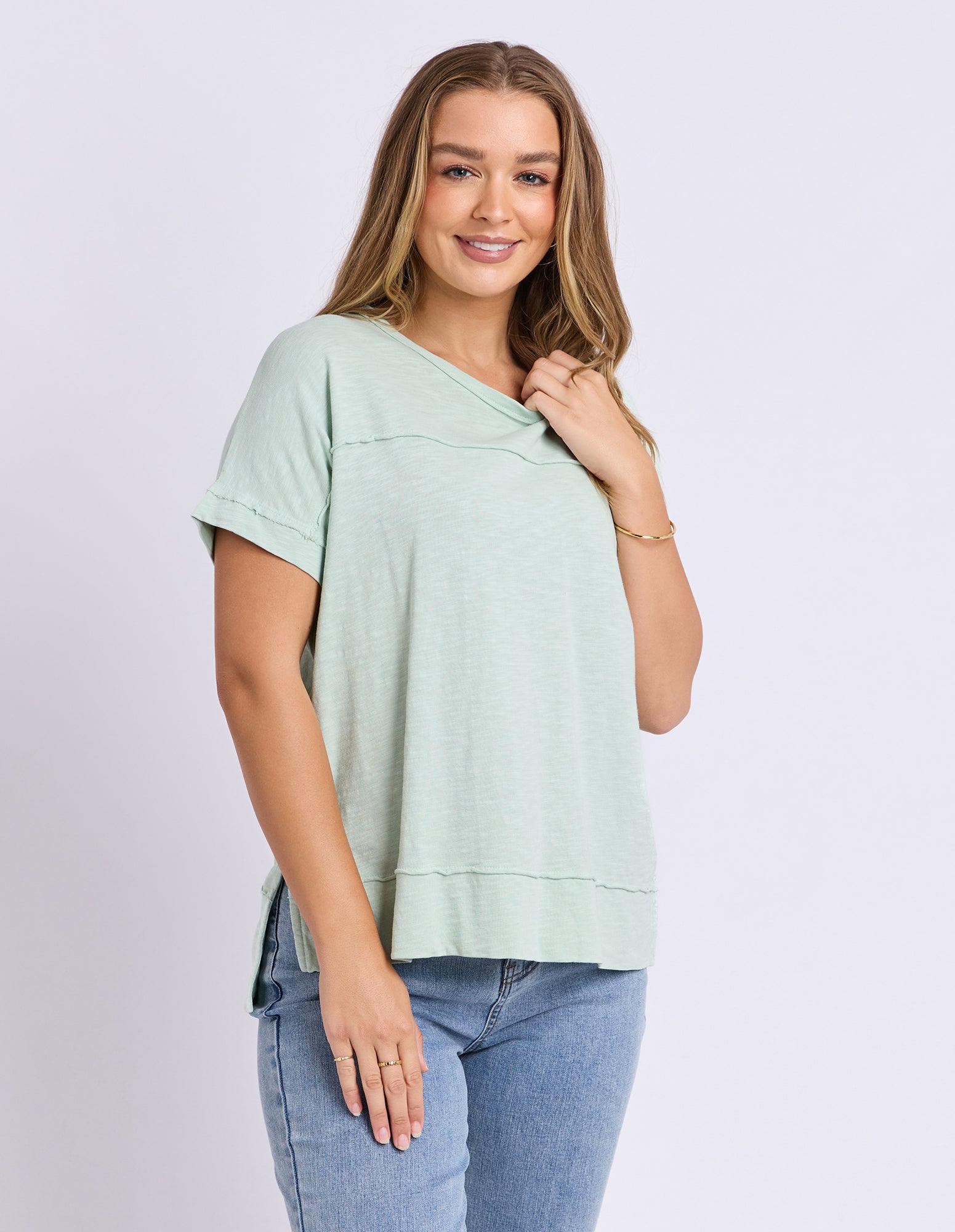 A woman modeling a light sage green short-sleeve top with a split neckline, paired with blue jeans, against a plain light gray studio background.