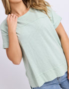 A woman wearing a light sage green short-sleeve t-shirt with a relaxed, slightly oversized fit and a raw, scalloped hem, paired with blue jeans against a light gray studio background.