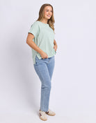 A woman modeling a light mint green short-sleeve t-shirt and light blue jeans, standing against a plain light gray studio background.