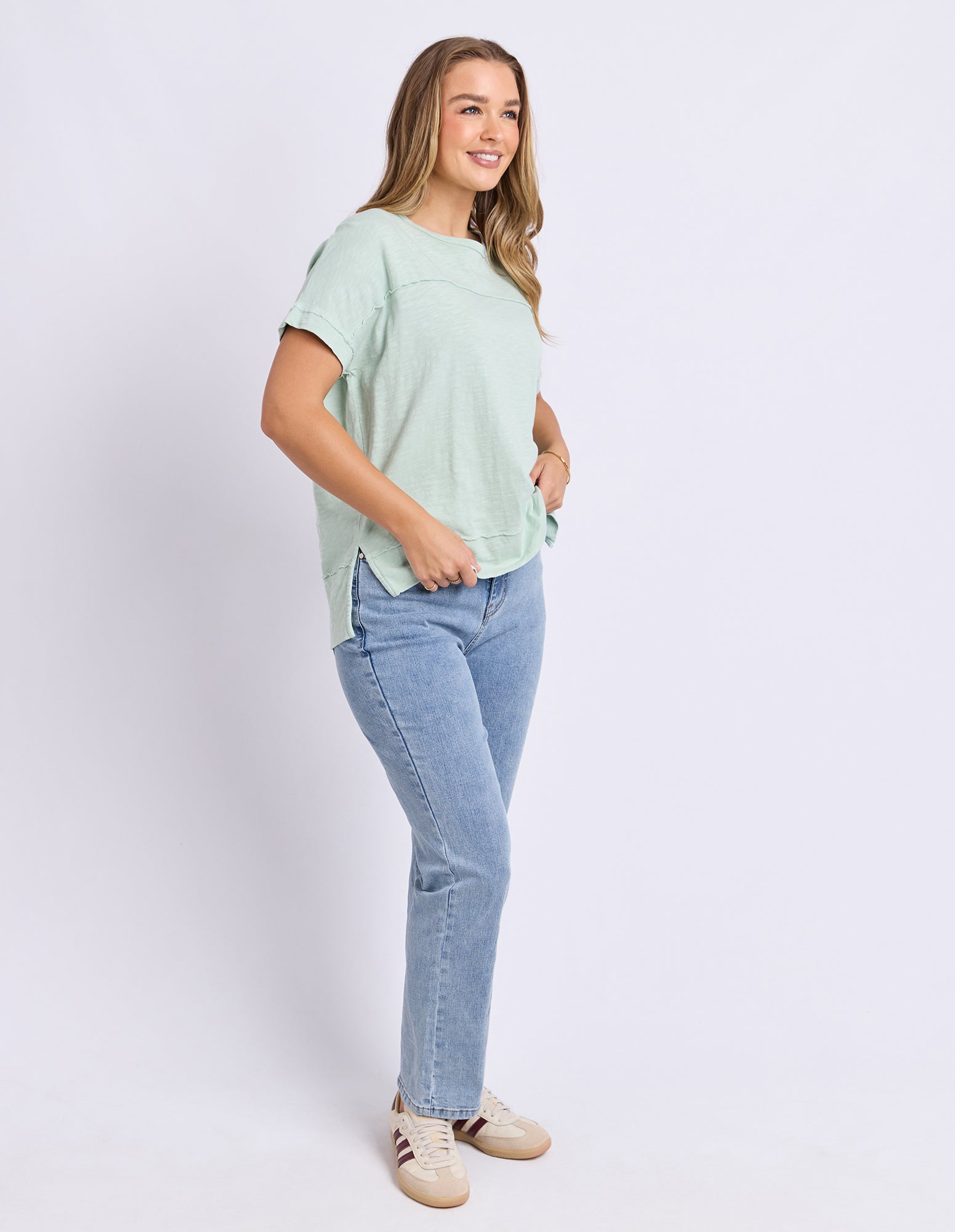 A woman modeling a light mint green short-sleeve t-shirt and light blue jeans, standing against a plain light gray studio background.