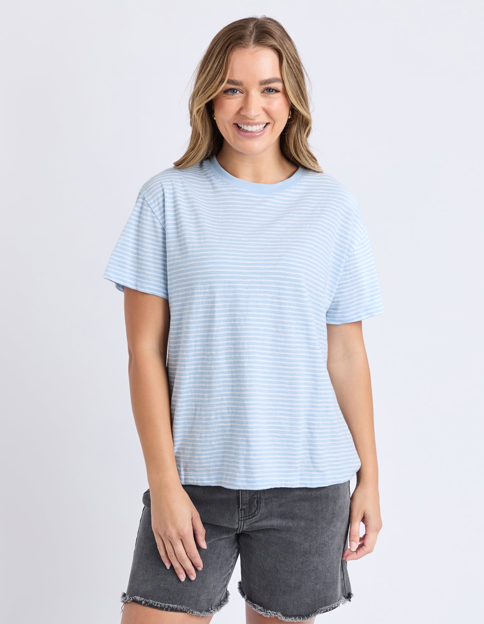 A woman modeling a light blue and white horizontally striped short-sleeve t-shirt with dark gray denim shorts, set against a plain light gray studio background.