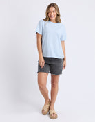 A woman modeling a light blue and white striped short-sleeve t-shirt, dark gray denim shorts, and beige platform sandals against a plain white studio background.