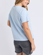A woman wearing a light blue and white striped short-sleeve t-shirt and dark gray denim shorts, shown from a three-quarters back view against a light gray studio background.