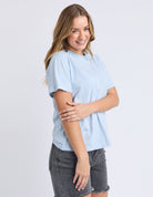A woman modeling a light blue and white striped short-sleeve t-shirt with dark gray denim shorts, standing against a light gray studio background.