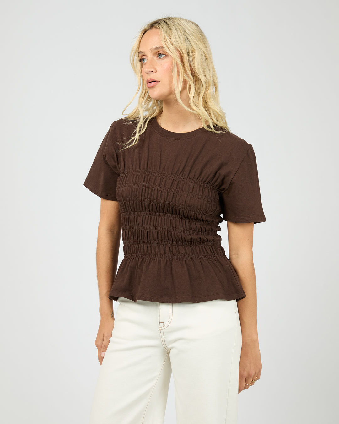 Woman wearing a brown ruched top and white pants on a plain background