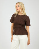 Woman wearing a brown ruched top and white pants on a plain background
