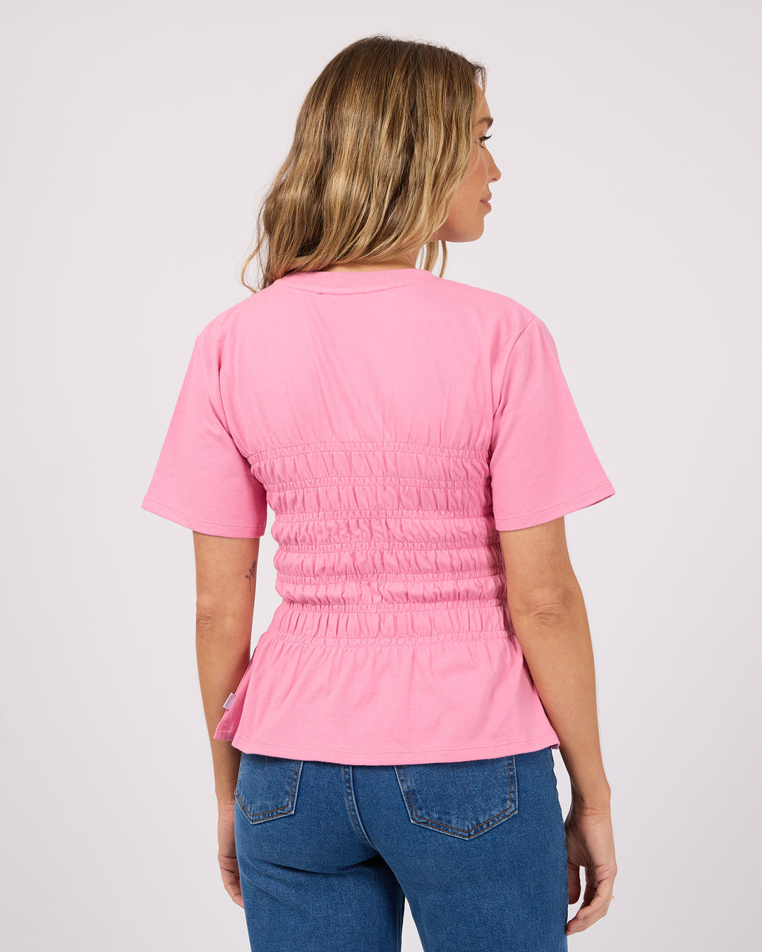 A woman viewed from the back wearing a pink short-sleeve scrub top with a smocked back panel and peplum hem, paired with blue jeans, against a light gray studio background.