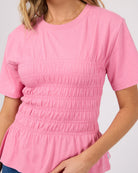 A close-up of a woman wearing a pink short-sleeve top with horizontal smocked gathering details across the chest and waist, paired with blue jeans against a light gray background.