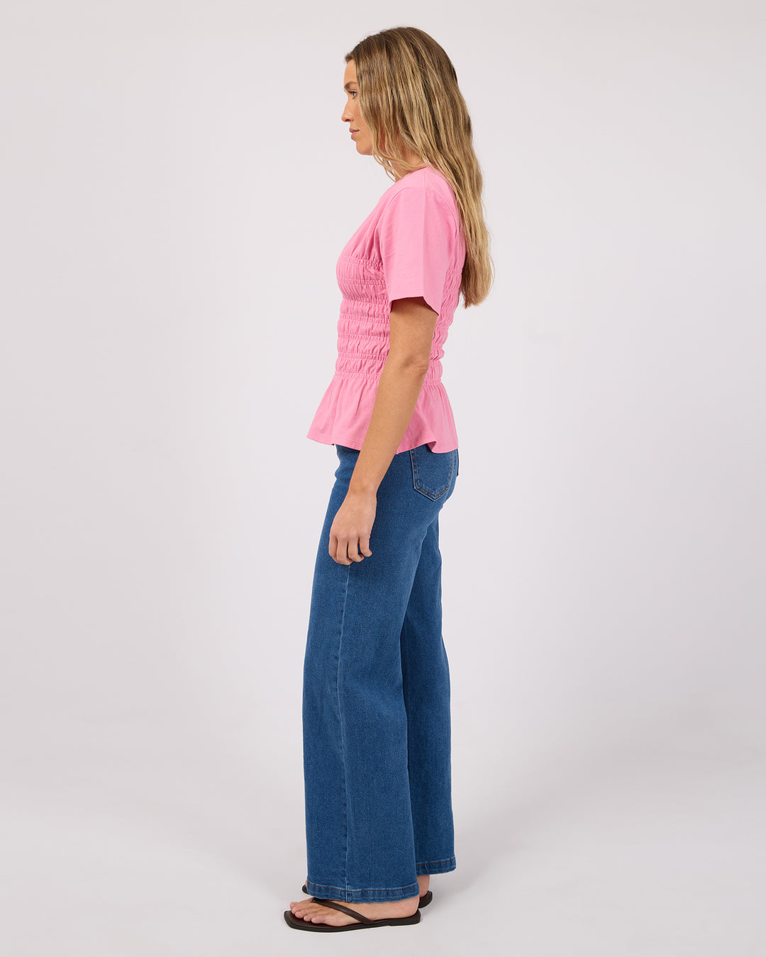 A woman stands in profile wearing a pink short-sleeve smocked blouse and blue wide-leg jeans against a plain light gray studio background.