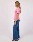 A woman stands in profile wearing a pink short-sleeve smocked blouse and blue wide-leg jeans against a plain light gray studio background.