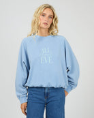 A blonde woman wearing a light blue crewneck sweatshirt with 'ALL about EVE' printed in light blue on the front, paired with blue jeans, against a light gray studio background.