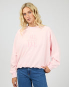 A blonde woman wearing a light pink crewneck sweatshirt with the words 'ALL night LOVE' printed in tonal pink on the front, paired with blue jeans, against a light gray studio background.