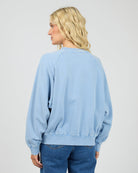A woman with blonde hair is shown from the back wearing a light blue crewneck sweatshirt and blue jeans against a plain light gray background.