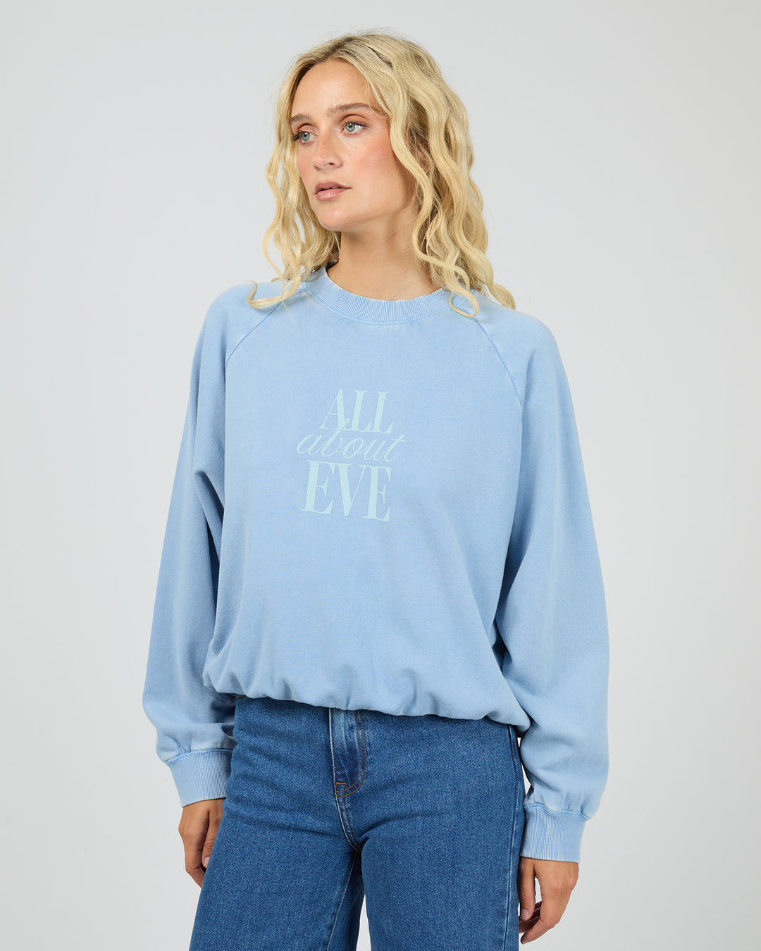 A woman wearing a light blue crewneck sweatshirt with 'ALL ABOUT EVE' printed in a matching pale blue script on the front, paired with blue jeans, against a light gray studio background.