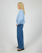 A woman stands in profile wearing a light blue long-sleeve blouse and wide-leg blue jeans against a plain light gray studio background.
