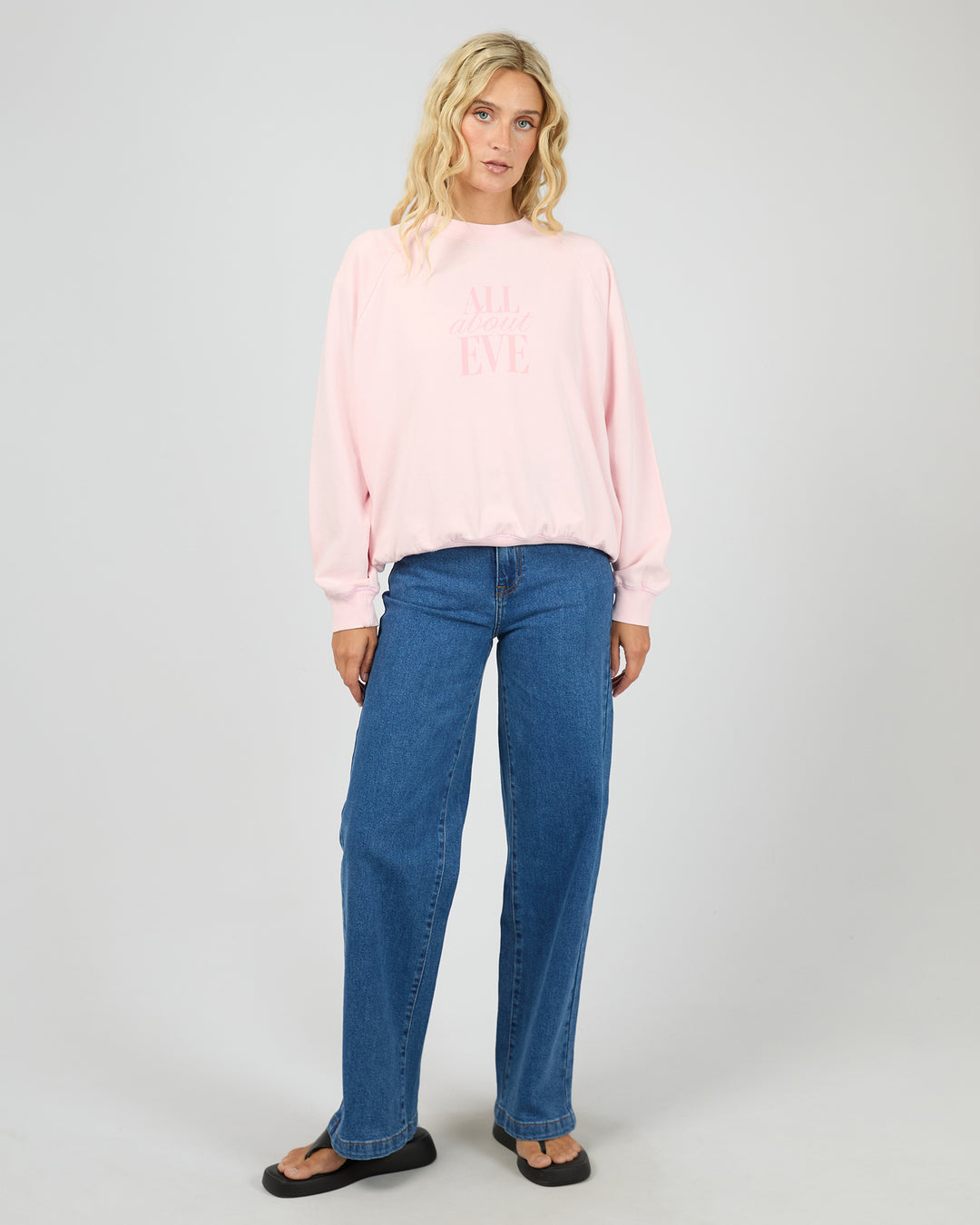 A woman modeling a light pink crewneck sweatshirt with pink text on the chest, paired with blue jeans and black sandals, against a light gray studio background.