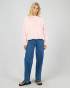 A woman modeling a light pink crewneck sweatshirt with pink text on the chest, paired with blue jeans and black sandals, against a light gray studio background.