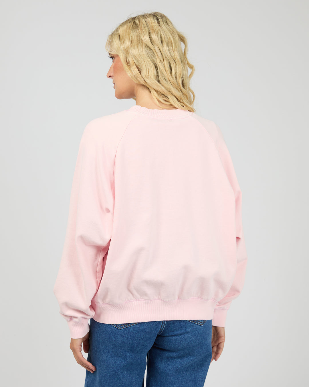 A woman with blonde hair is shown from the back wearing a light pink crewneck sweatshirt and blue jeans against a light gray studio background.