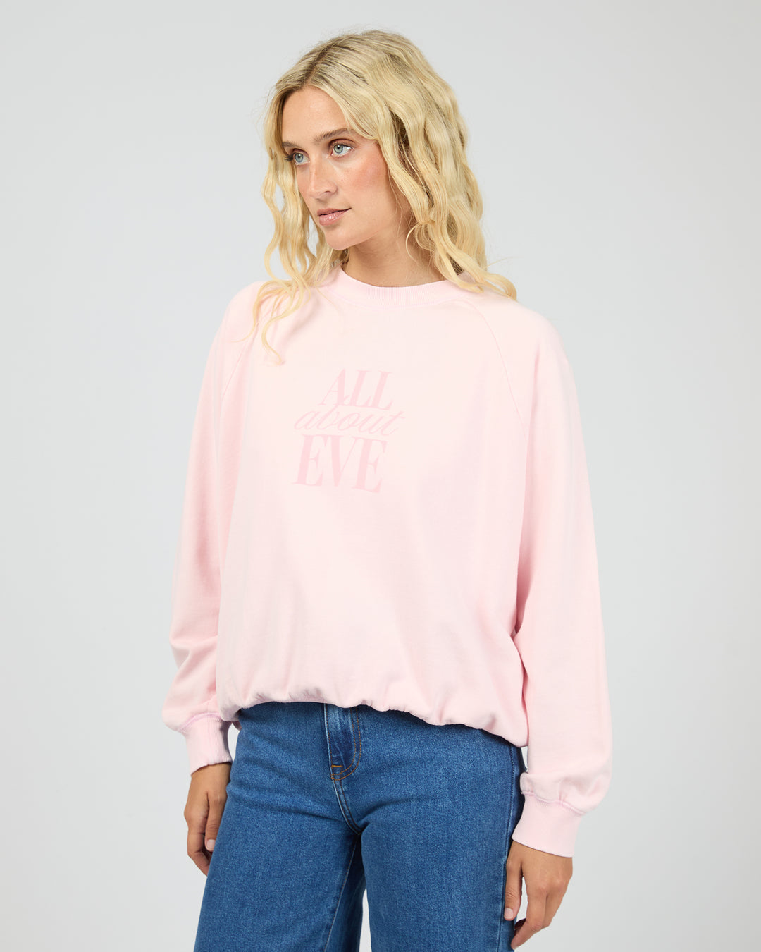 A blonde woman wearing a light pink crewneck sweatshirt with 'ALL about EVE' printed in tonal pink text on the front, paired with blue jeans, against a light gray studio background.