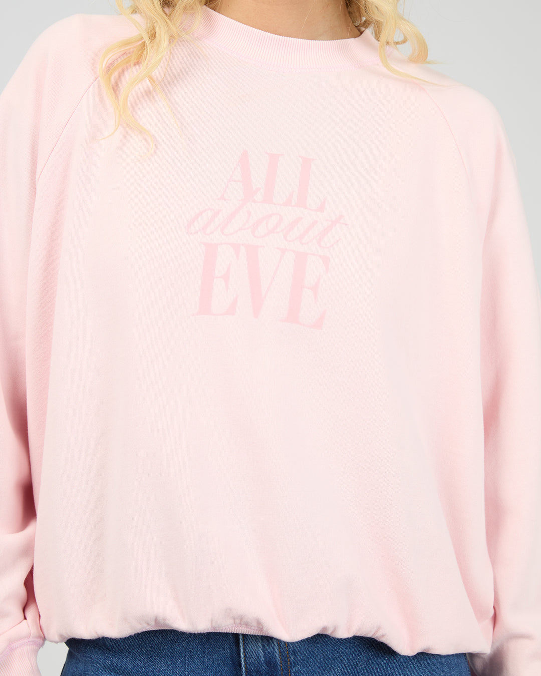 A close-up of a person wearing a light pink crewneck sweatshirt with the words 'ALL about EVE' printed in a matching pale pink tone on the front, paired with blue jeans.