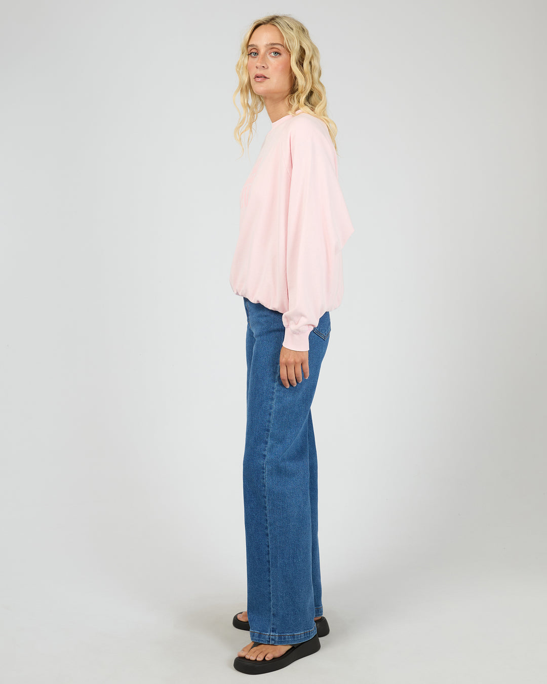 A woman modeling a light pink long-sleeve top and blue wide-leg jeans against a plain light gray studio background.