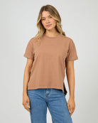A woman modeling a cropped, short-sleeve crewneck t-shirt in a washed, muted brownish-pink color, paired with blue jeans, against a light gray studio background.