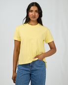 A woman modeling a pale yellow short-sleeve crewneck t-shirt paired with blue jeans, standing against a light gray studio background.