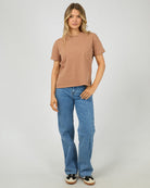A woman modeling a cropped, short-sleeve crewneck t-shirt in a washed tan color, paired with blue jeans and black-and-white sneakers, against a light gray studio background.
