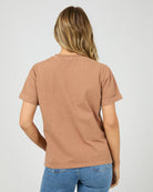 A woman viewed from behind wearing a muted brown short-sleeve t-shirt with rolled cuffs and blue jeans, set against a plain light gray studio background.