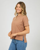 A woman wearing a short-sleeve crewneck t-shirt in a dusty rose-brown color, paired with blue jeans, photographed against a solid light gray background.