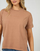A woman wearing a muted dusty rose-brown short-sleeve t-shirt with 'ALL ABOUT ME' printed in tonal embroidery on the chest, paired with blue jeans against a light gray background.
