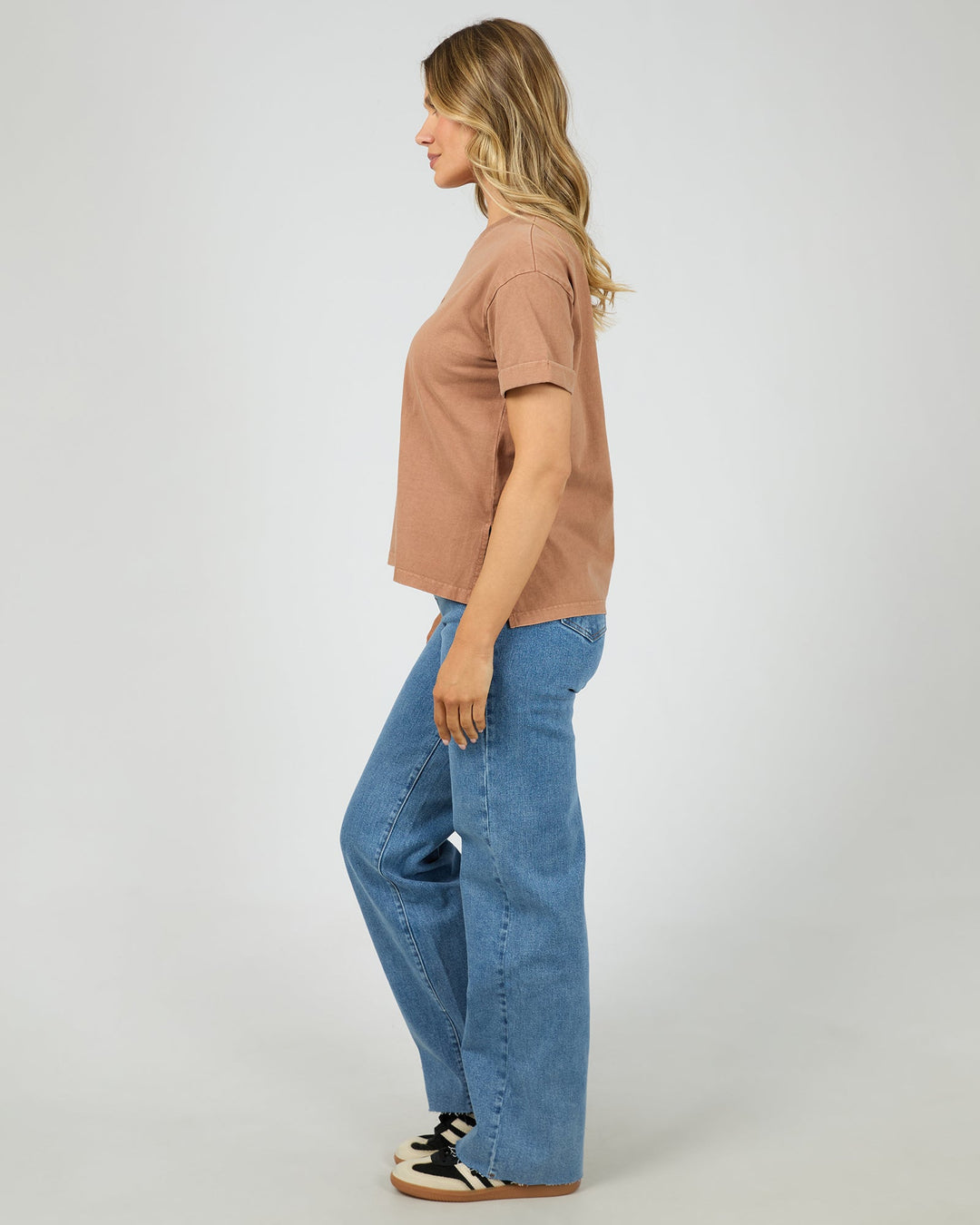 A woman shown in profile wearing a cropped, short-sleeve dusty rose t-shirt, wide-leg blue jeans, and black-and-white sneakers against a light gray studio background.