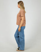 A woman shown in profile wearing a cropped, short-sleeve dusty rose t-shirt, wide-leg blue jeans, and black-and-white sneakers against a light gray studio background.