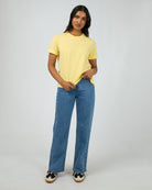 A woman modeling a pale yellow short-sleeve crewneck t-shirt and wide-leg blue jeans against a light gray studio background.