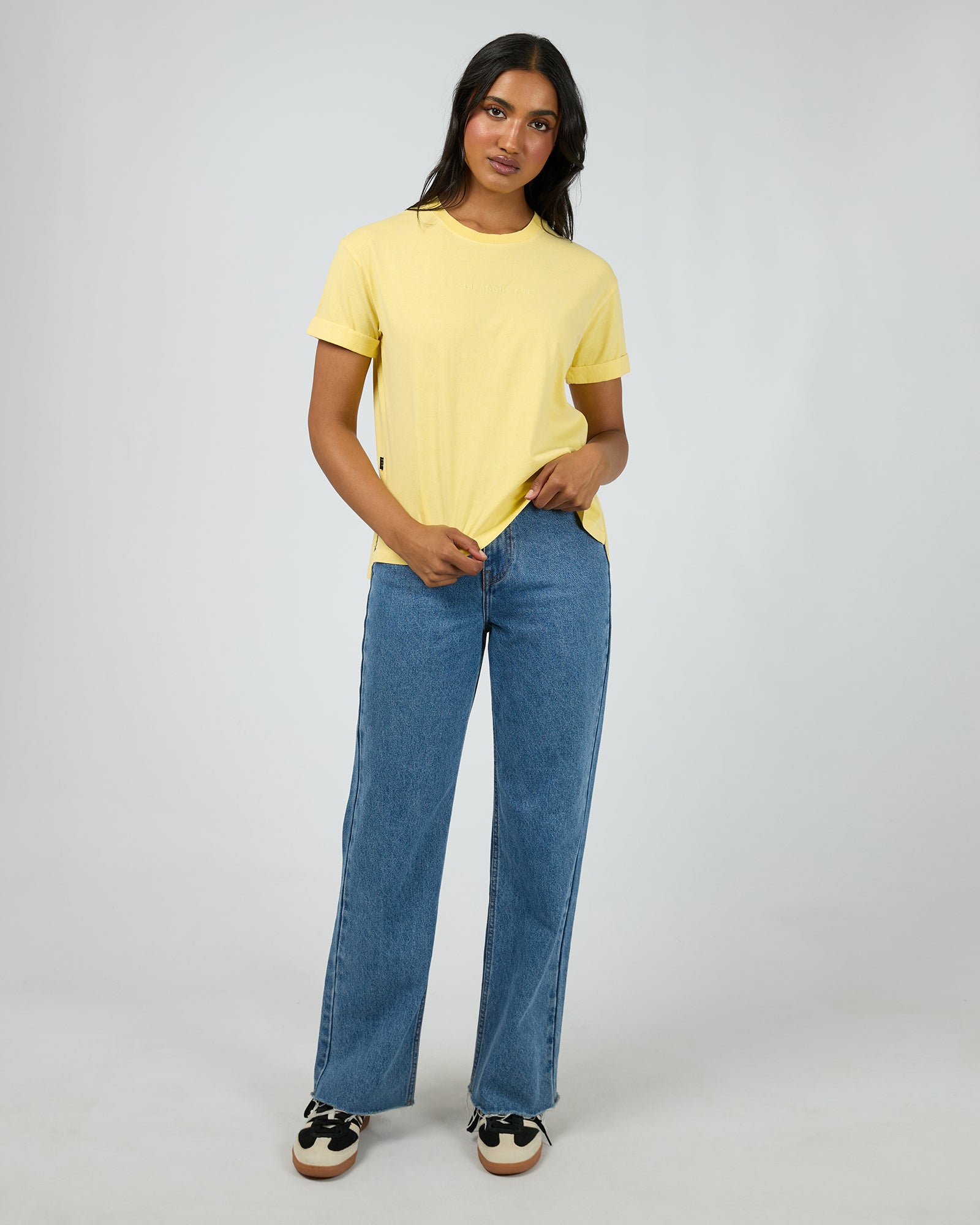 A woman modeling a pale yellow short-sleeve crewneck t-shirt and wide-leg blue jeans against a light gray studio background.